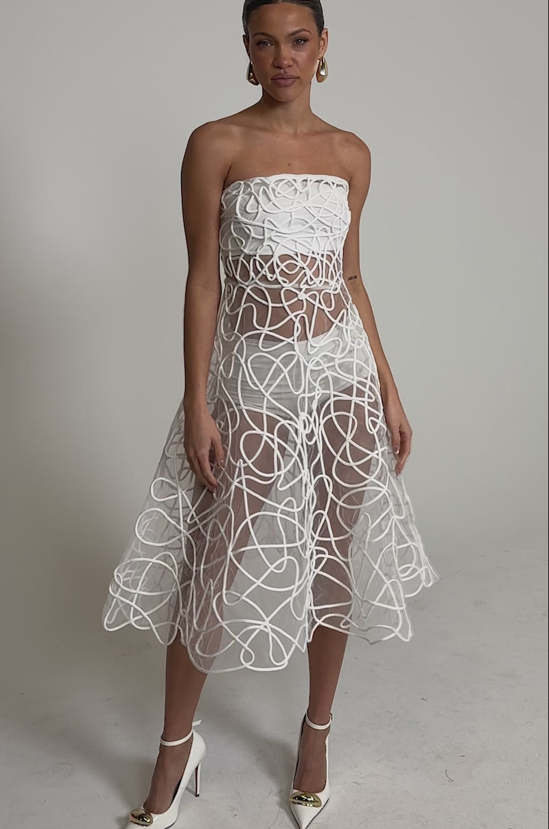 A video of a woman wearing the MAYA ABSTRACT MIDI DRESS IN WHITE—a strapless sheer midi with swirling patterns—styled with gold pointed-toe heels and hoop earrings, standing against a plain background, perfect for cocktail parties.