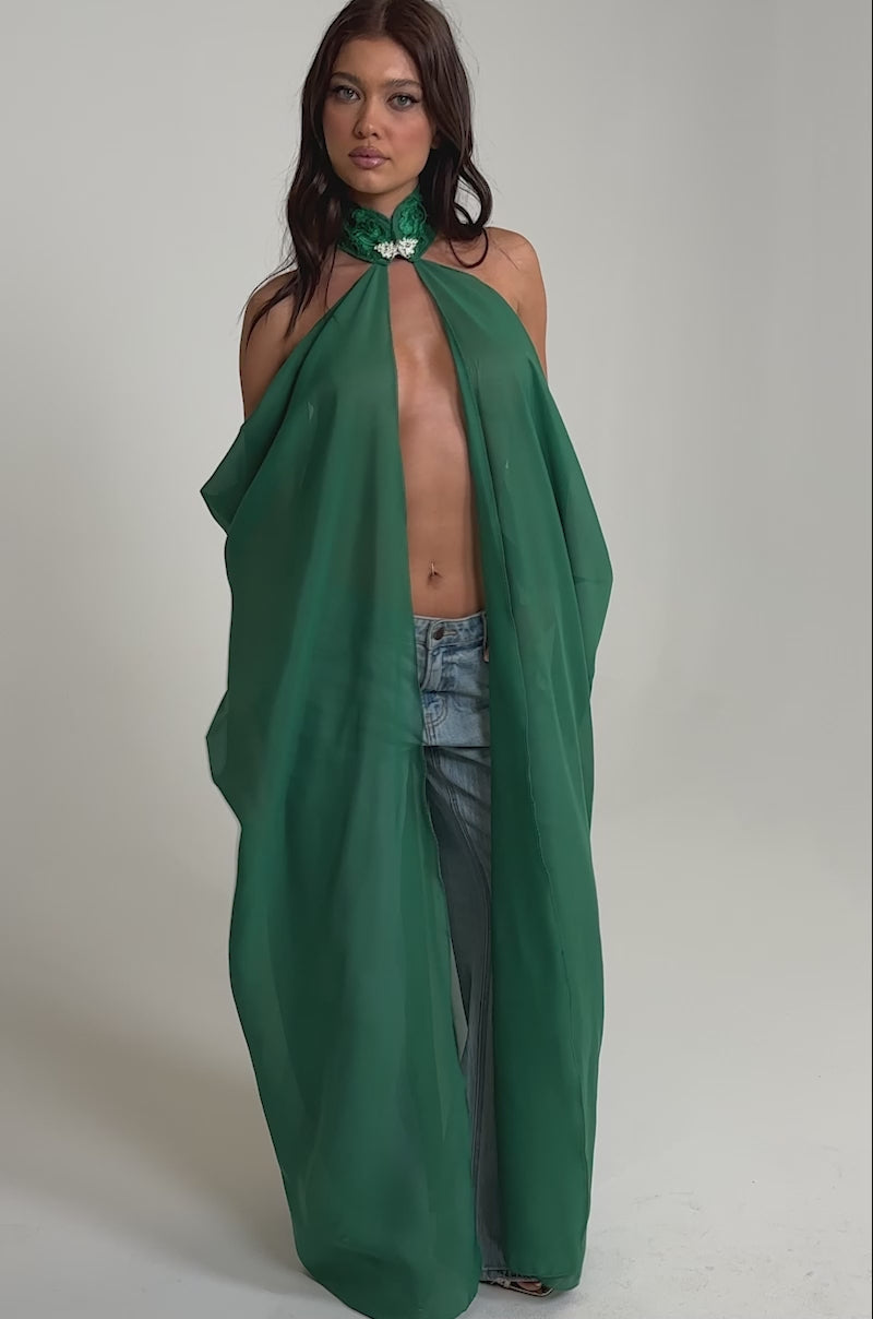 Video of woman modeling the MAKE A STATEMENT CAPE IN GREEN, a sheer green halter-style cape with embellished collar and floral detail. The cape is open at the front, revealing blue jeans. She has long, wavy hair and a neutral expression.