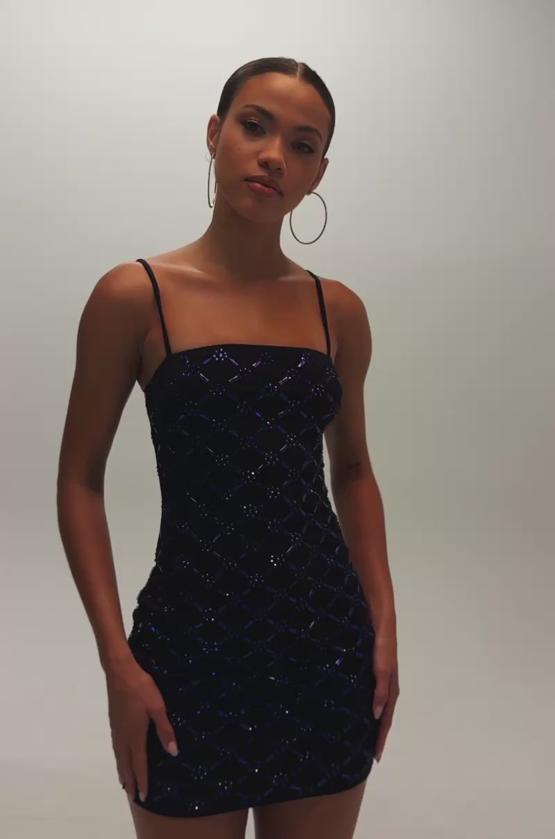 Video of woman modeling the SPARKLE CITY RHINESTONE MINI DRESS IN NAVY, a fitted sleeveless mini dress with thin straps and a striking blue and black geometric pattern. She accessorizes with hoop earrings and has her hair pulled back.