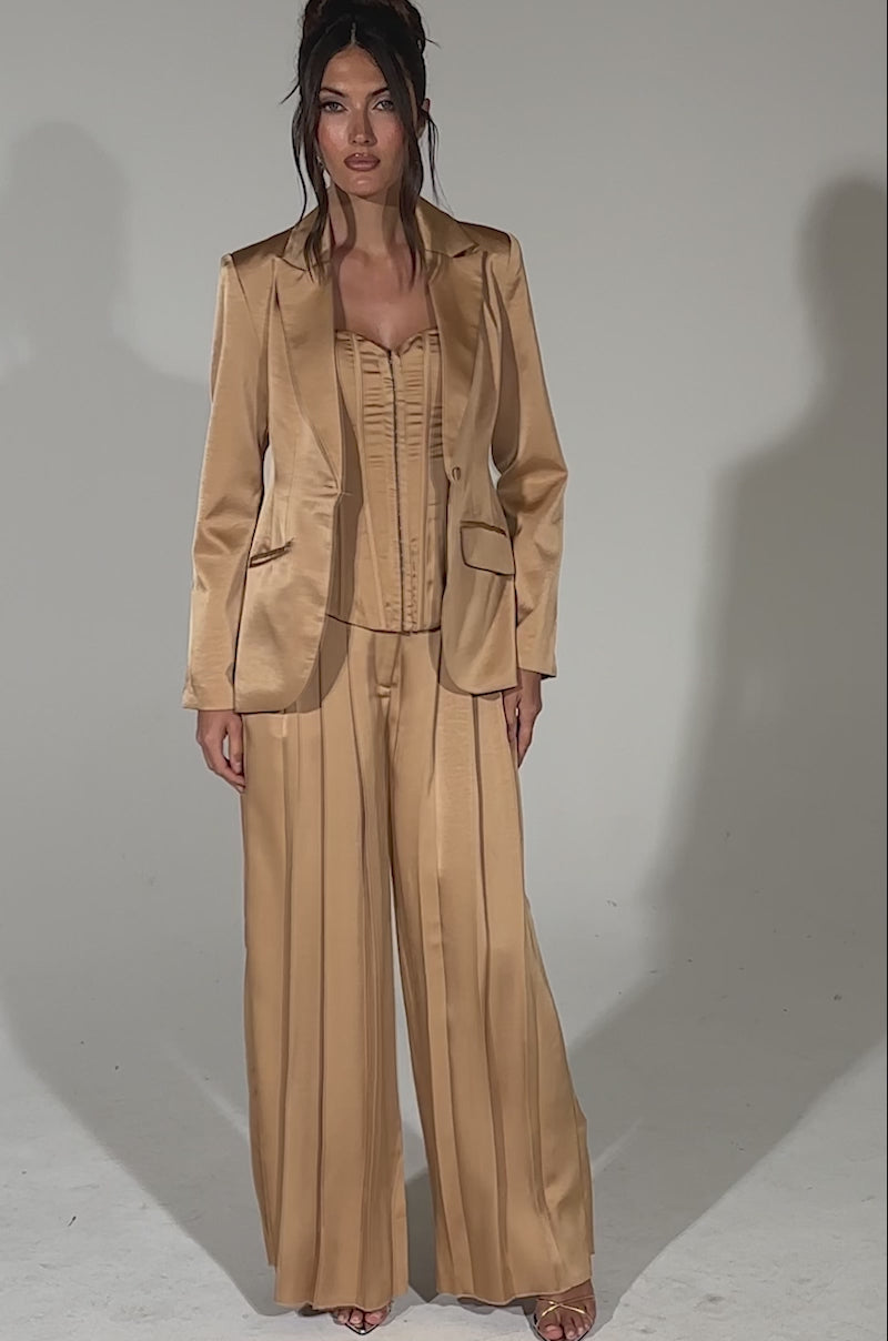 A video of a woman standing against a light background wearing HANSEL MOCHA SATIN PALAZZO PANT with a fitted corset top and tailored blazer. She has long, wavy hair styled in a loose updo and pairs the look with open-toe heels.