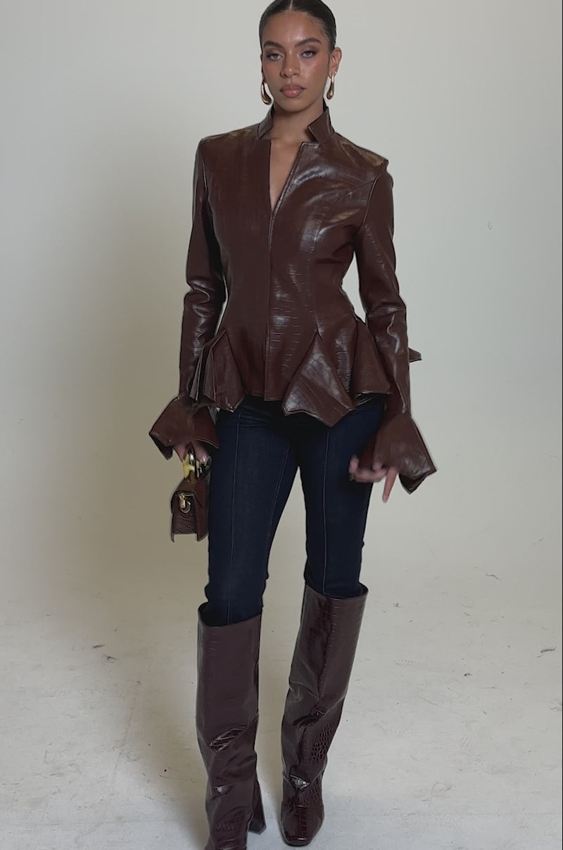 A video of a woman in the CARMEN BODYCON BLAZER IN BURGUNDY, styled with dark blue jeans, tall brown crocodile-textured boots, and a small brown handbag against a light gray background.