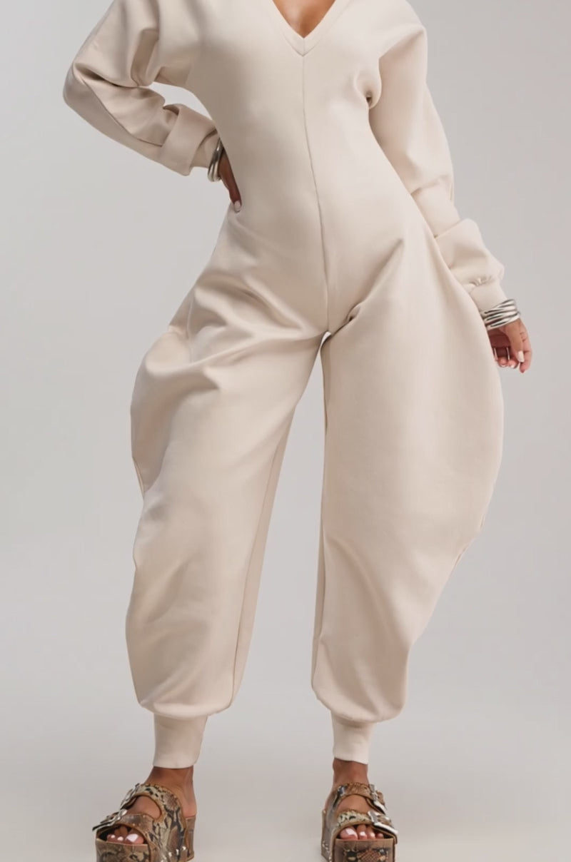 Video of woman modeling the COMFY COZY SWEATSHIRT JUMPSUIT IN IVORY, featuring exaggerated rounded hips and a V-neckline. Her hair is in a bun, styled with platform sandals and bracelets, set against a plain light background.