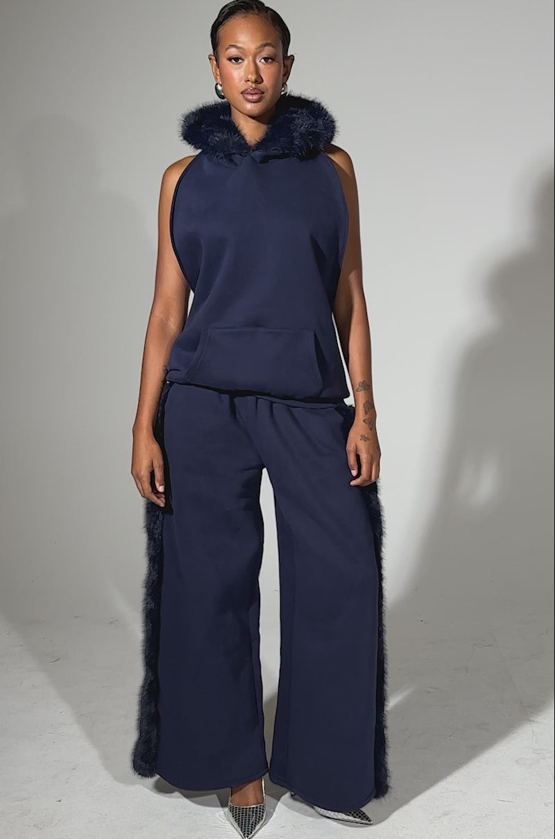 A video of a woman modeling the LUXE FUR OPEN BACK SWEATSHIRT IN NAVY, a sleeveless sweatshirt with a fur-trimmed hood and matching wide-leg pants, styled with pointed silver heels against a light blue background.