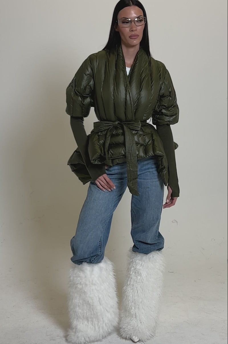A video of a woman standing against a plain background, wearing clear goggles, blue jeans, fluffy white boots, and the RIB SLEEVE PEPLUM PUFFER IN OLIVE.
