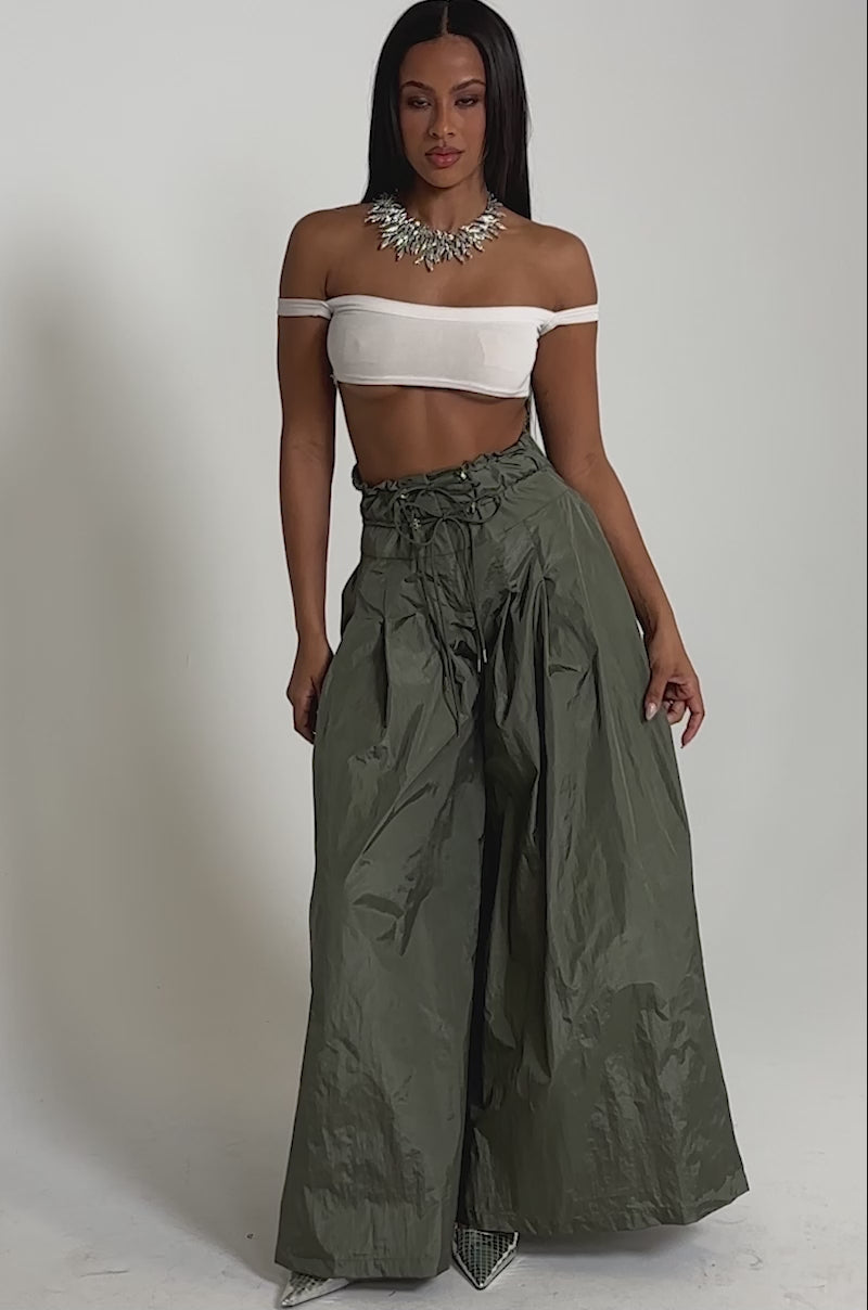A video of a woman in an off-the-shoulder white crop top, DANCE THE NIGHT AWAY PALAZZO PANT in olive green with a high-rise drawstring waist, silver pointy heels, and a statement necklace on a plain background.