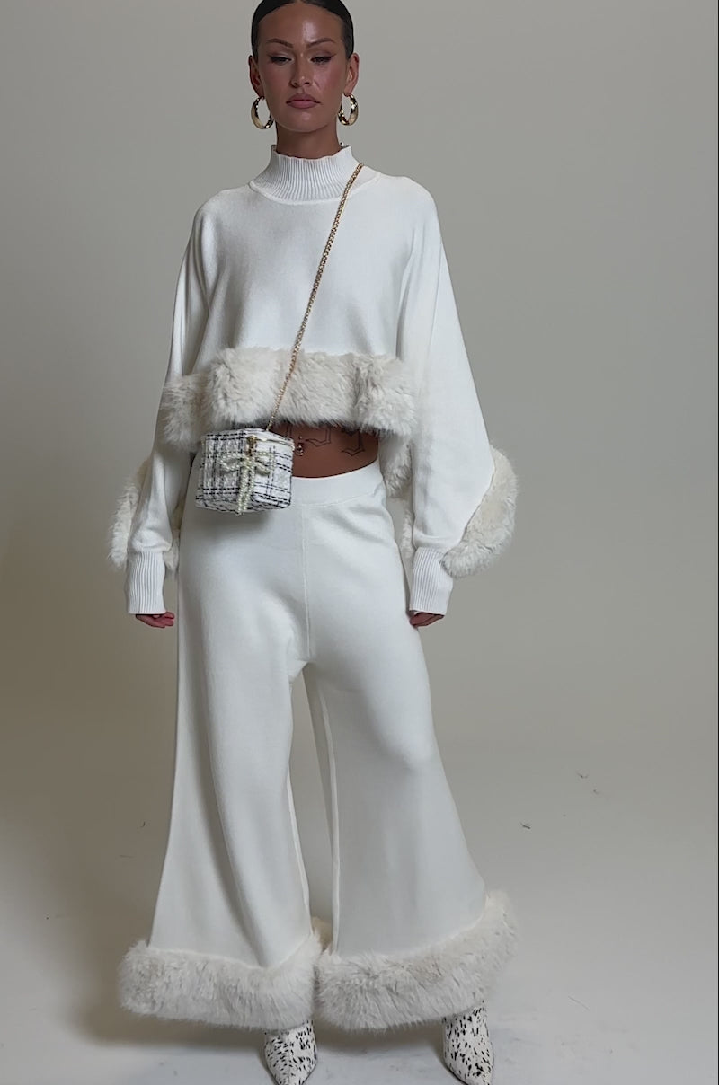 A video of a woman in the QUIET LUX FUR TRIM SWEATER PANTS IN WHITE, styled with a matching long-sleeve crop top, gold hoop earrings, and white patterned boots for a chic knit look.