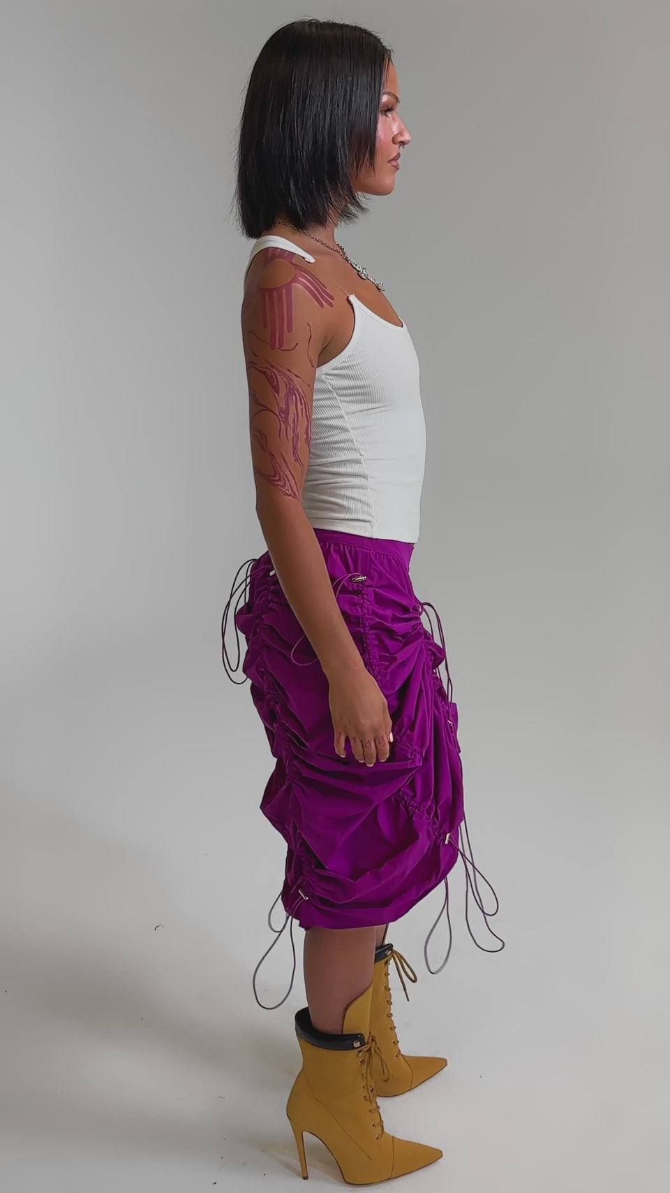 A video of a woman wearing a white tank top, the BELLA PARACHUTE MAXI SKIRT IN FUCHSIA, and yellow lace-up boots as she stands confidently against a plain light background.