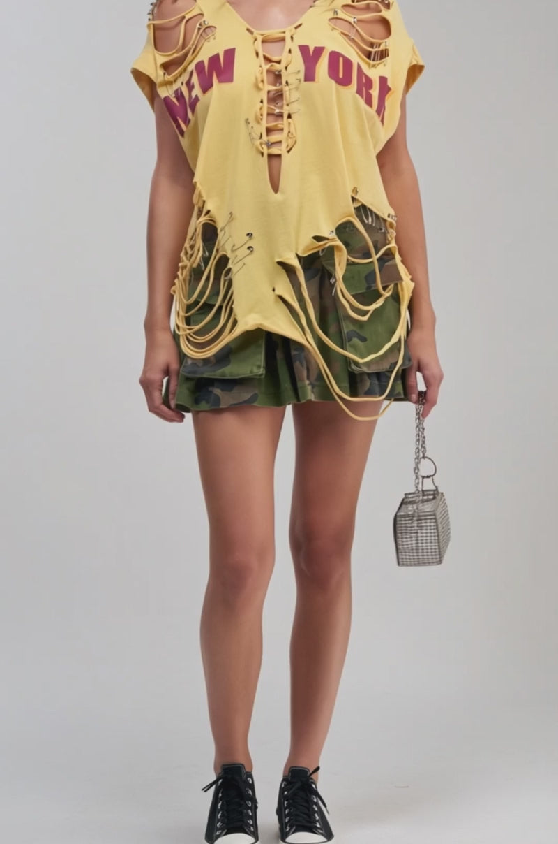 Video of a woman modeling the VARSITY STAR DISTRESSED TEE, a yellow graphic top adorned with a bold “New York” design, laces, and safety pins. 