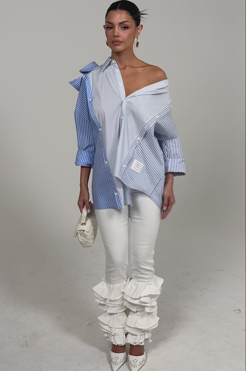 Video of woman modeling the ALL OVER THE PLACE BUTTON DOWN—an oversized, striped blouse with an asymmetrical layered design—with white pants and a textured cream clutch. Her hair is pulled back as she poses against a plain background.