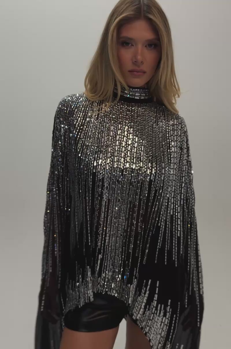 Video of a woman modeling the DRIPPING IN RHINESTONES CAPE, a glamorous black mesh piece with floor-length sheer, silver-studded rows, paired with black shorts and knee-high black boots.