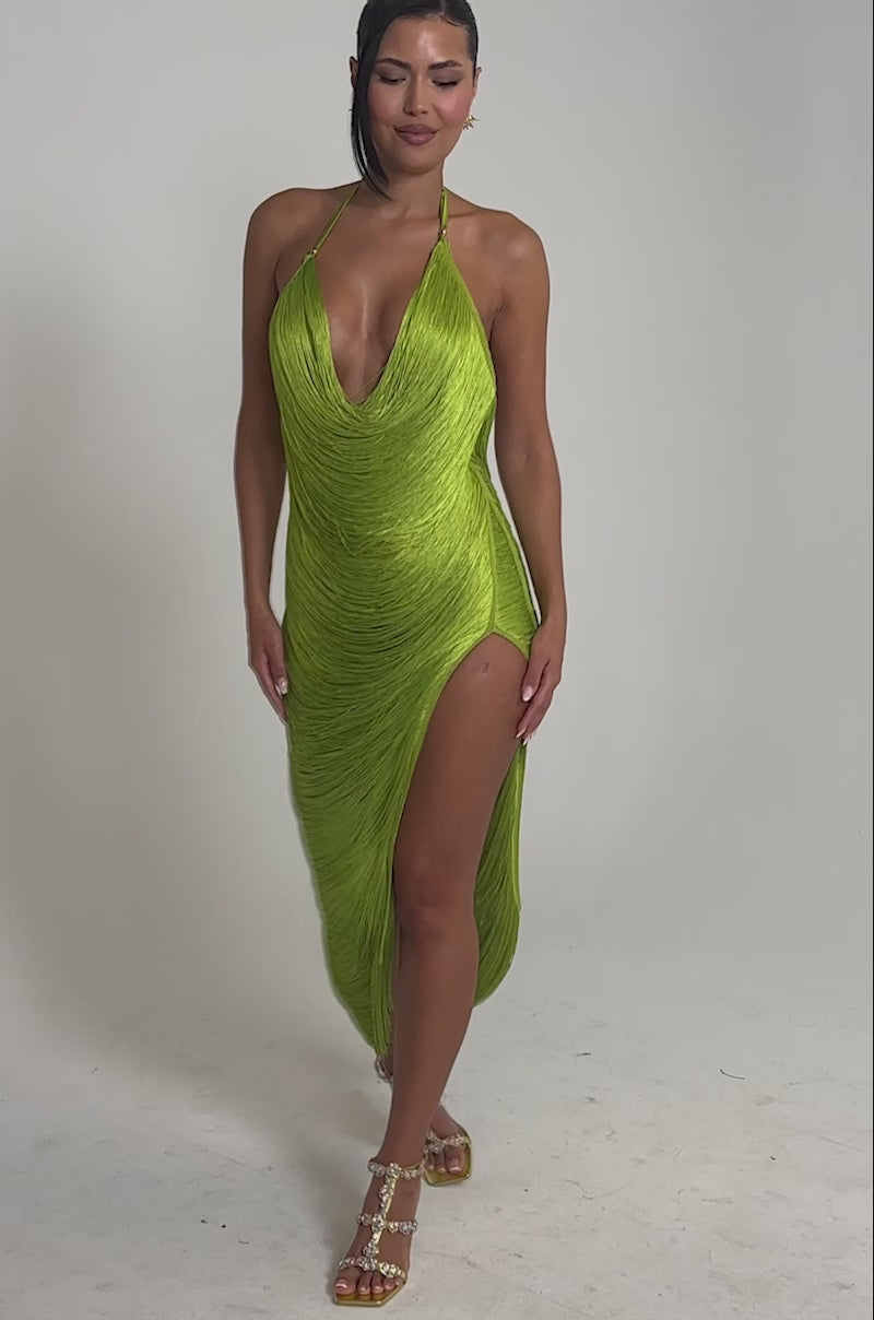 Video of woman modeling the SOFIA FRINGE HIGH SLIT MIDI DRESS IN GREEN, featuring a sleeveless design, deep neckline, fringe details, and high slit. She completes the bold look with strappy heels against a light gray background.
