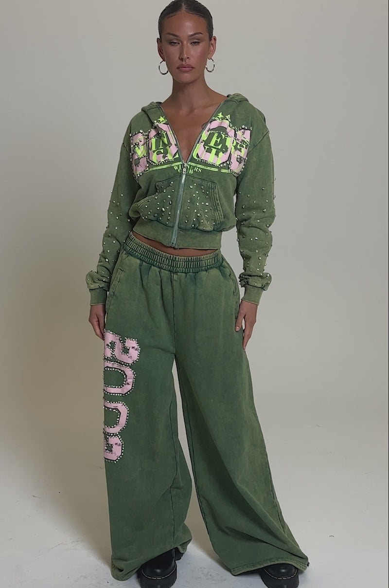 Video of woman modeling the WINNERS CLUB ZIP UP SWEATSHIRT in olive green with matching sweatpants. Pink and green graphics, rhinestone details, and a relaxed sporty vibe complete her confident look against a plain background.
