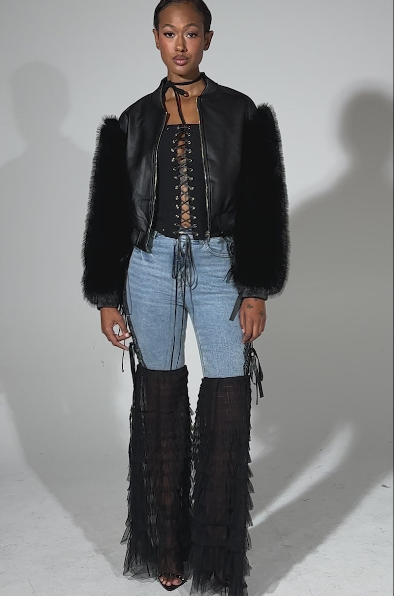 A video of a woman with short hair and layered necklaces facing forward, wearing the SO GOOD IT HURTS FUR SLEEVE BOMBER IN BLACK over a black lace bralette, high-waisted blue jeans, sheer ruffled black leg covers, and heeled sandals.