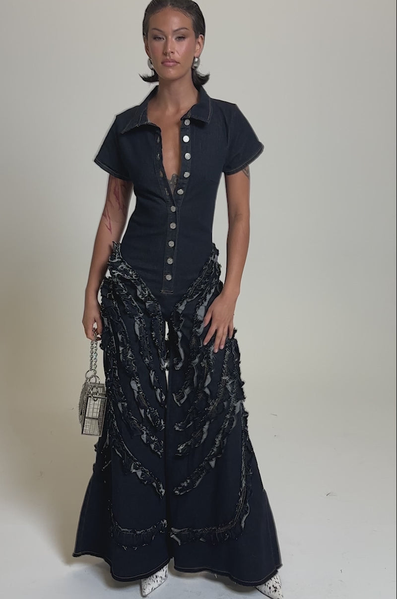 A video of a person in the INTO THE NIGHT DENIM JUMPSUIT, featuring dark wash denim with wide flared legs and frayed details, styled with a silver cage handbag and white boots against a light gray background.