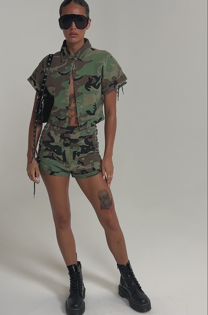 A video of a model wearing the I MEAN BUSINESS CAMO BUTTON DOWN and matching camo shorts, black combat boots, sunglasses, and a studded bag.