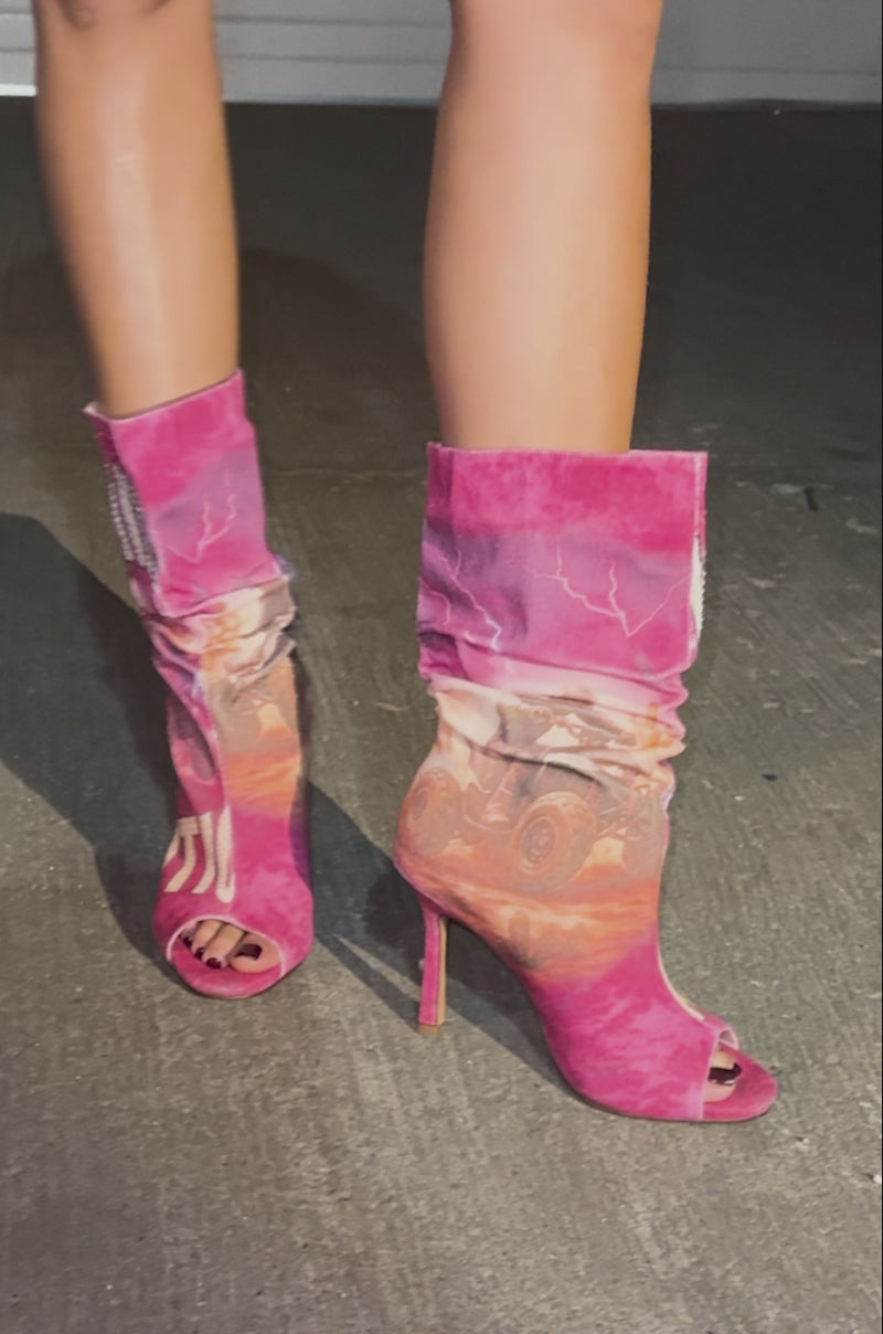 A video of a model wearing a sequined orange and green dress, standing on concrete at night in AKIRA GOLD FORMIDABLE PINK MULTI ABSTRACT OPEN TOE BOOTIES with stiletto heels and bold graphic text. Their face is not visible.