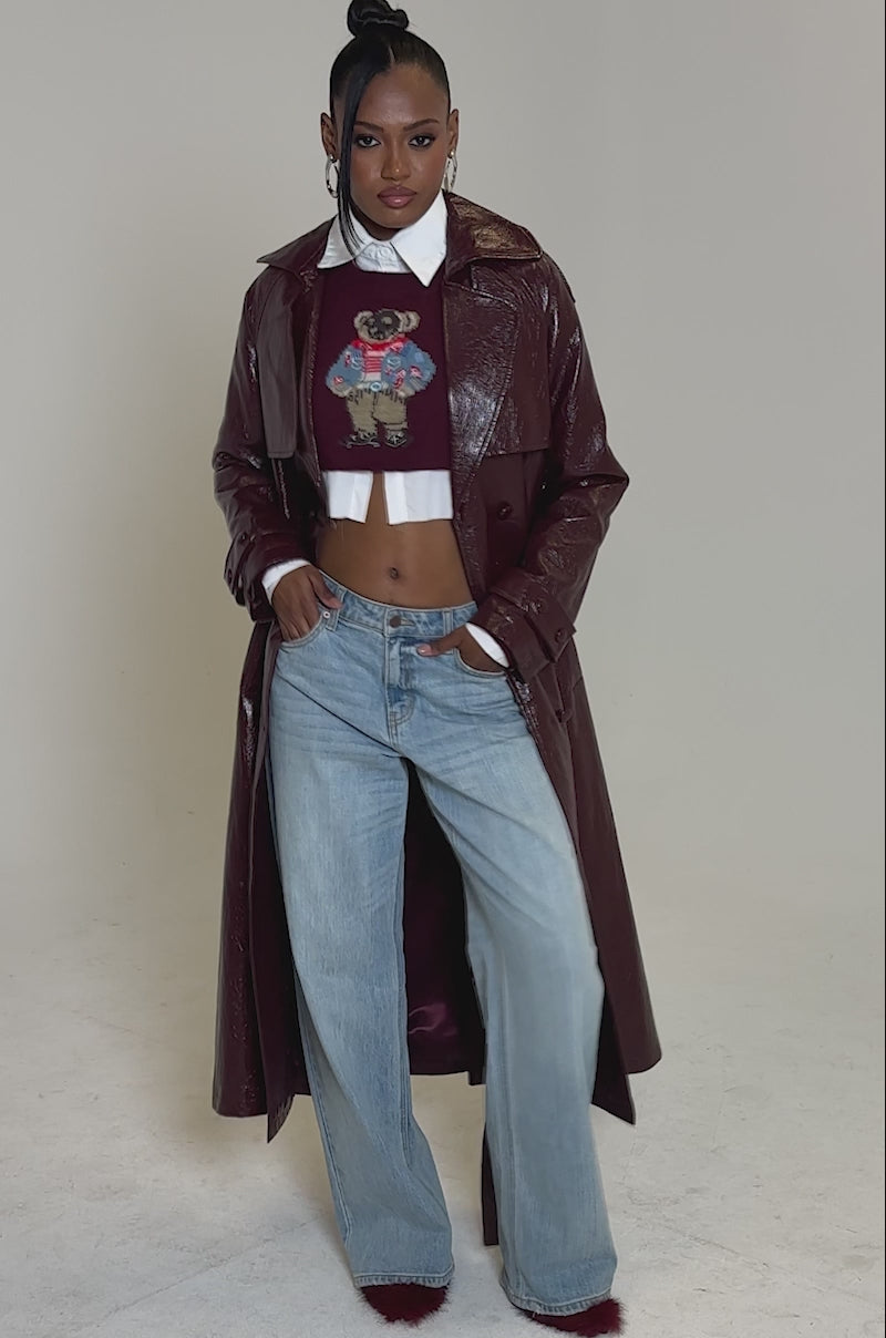 Video of woman modeling the WOKE UP IN PARIS FAUX LEATHER TRENCH, a shiny maroon longline coat, layered over a cartoon print crop top, wide-leg blue jeans, and red furry shoes. Her hair is styled in a high bun.