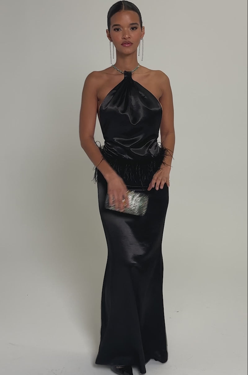 Video of woman modeling the WHO IS SHE SATIN FEATHER MAXI DRESS IN BLACK, featuring a feather-accented waist, and holds a small round silver clutch while standing against a plain light gray background.