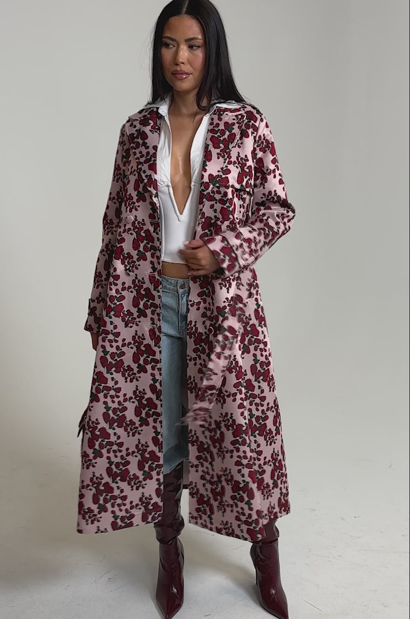 A video of a woman in the BUY ME ROSES JACQUARD TRENCH with a white corset top, blue jeans, knee-high burgundy boots, and a matching burgundy handbag. 