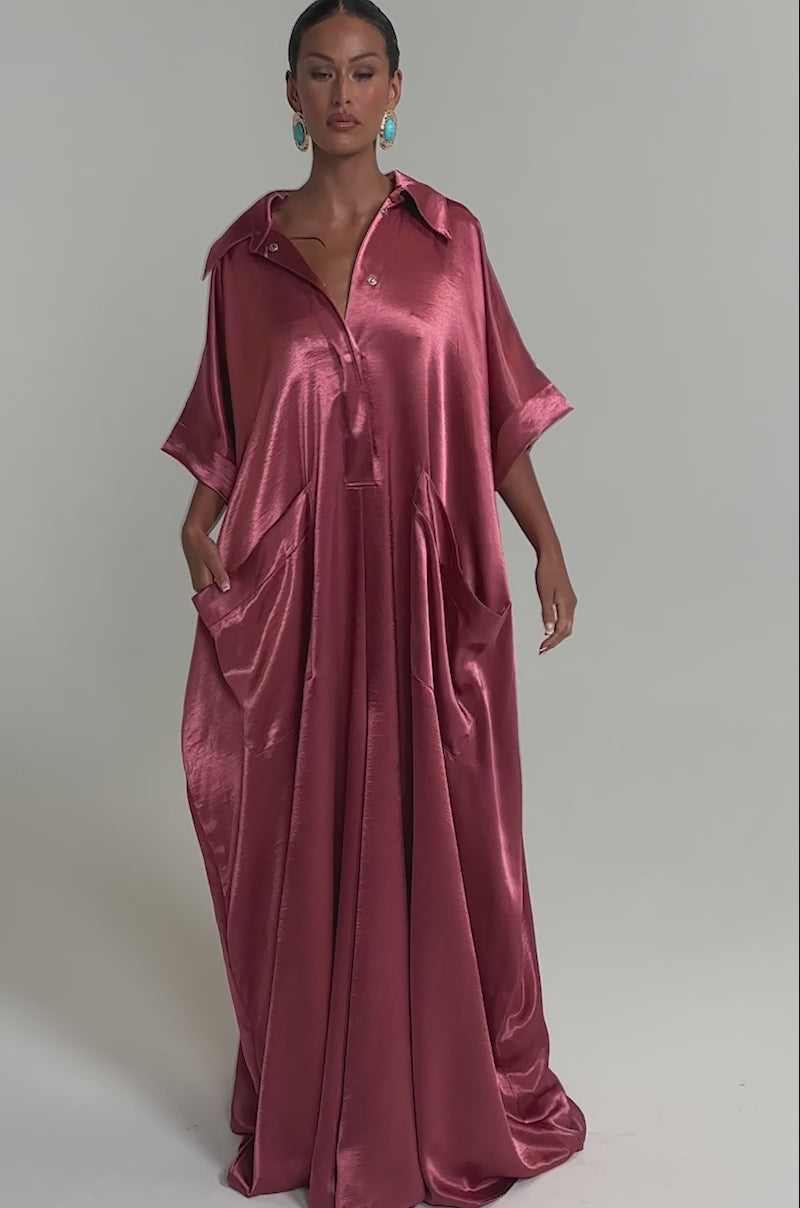 Video of woman modeling the SHE'S RICH OVERSIZED JUMPSUIT IN ROSE, a loose, satin pink wide-leg style with short sleeves, a collar, front buttons, and large pockets—styled with turquoise earrings for modern chic eveningwear.