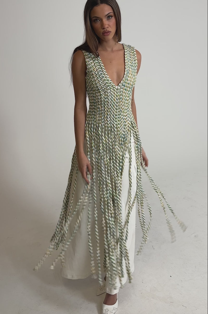 Video of woman modeling the TIED DOWN V-NECK ROPE TOP, a sleeveless floor-length top with a white base and green and yellow braided patterns, pairs it with white pointed-toe shoes. She has straight, shoulder-length brown hair.
