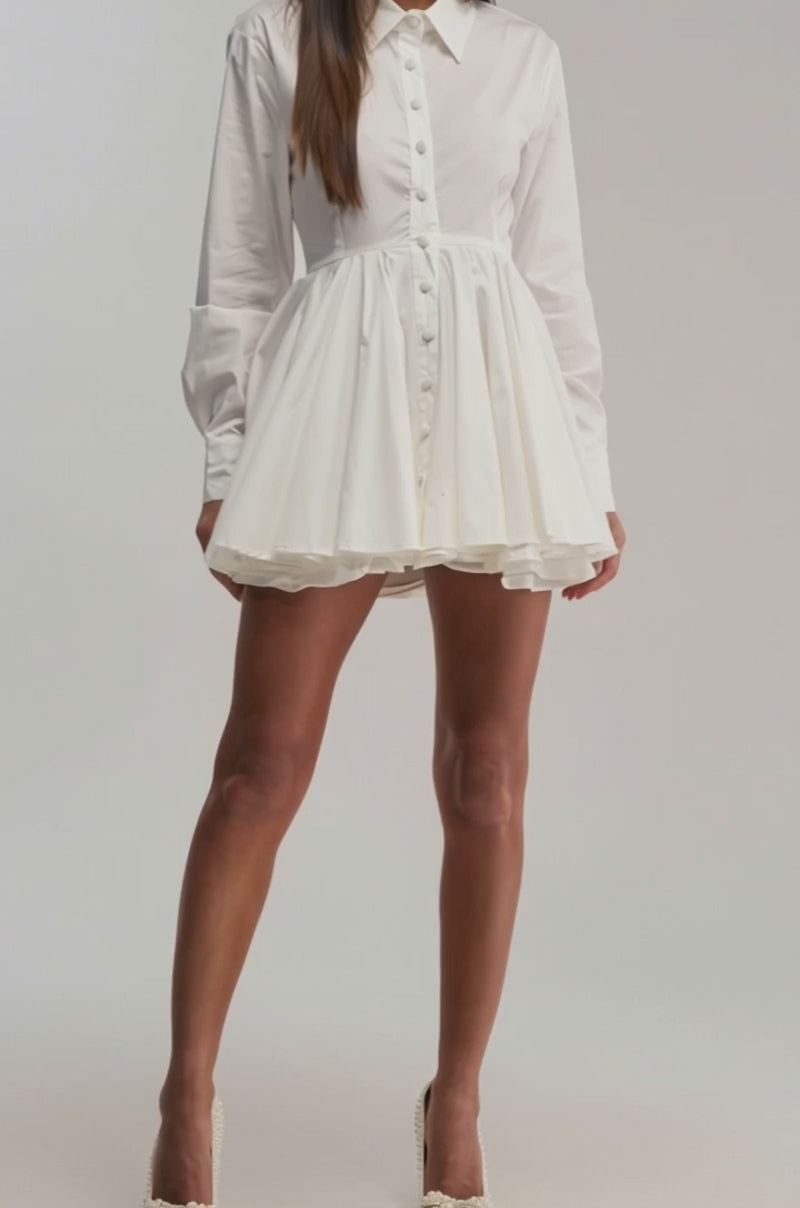 A video of a woman wearing THE IT GIRL POPLIN MINI DRESS—a chic white mini with long sleeves, a button-up front, and flared short skirt—paired with white heels. 