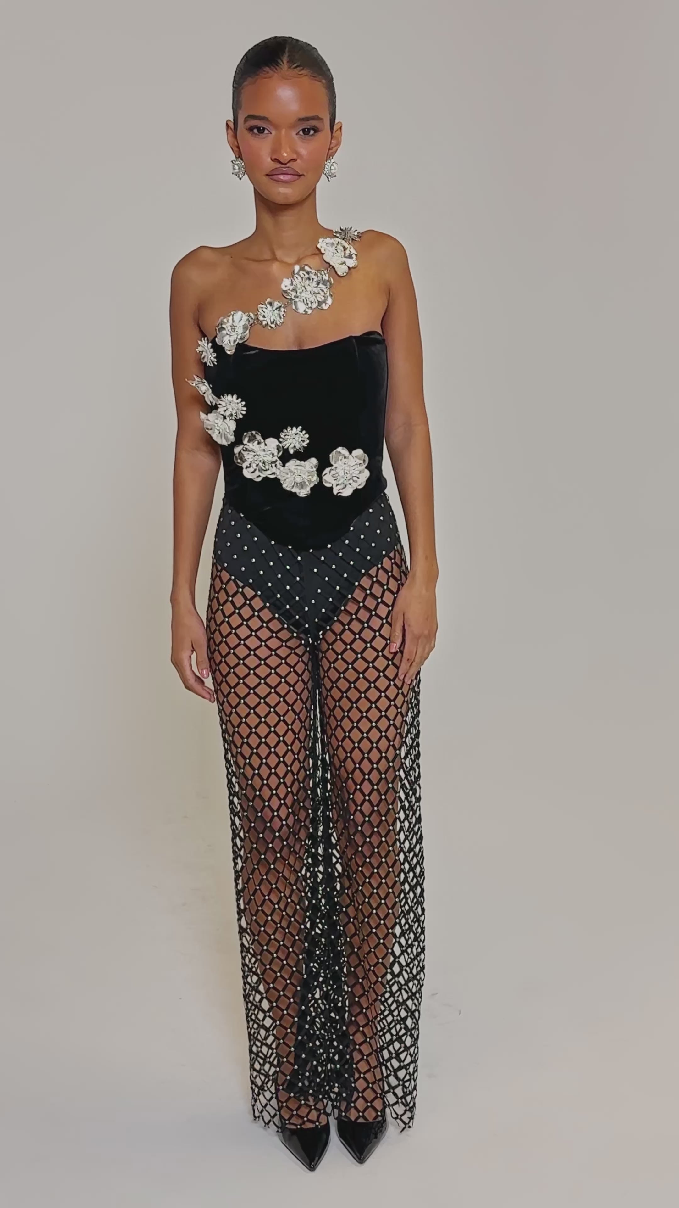 Video of woman modeling the CELINE STUD MESH PANT styled with a black, one-shoulder bodysuit featuring large white floral embellishments and black high heels.