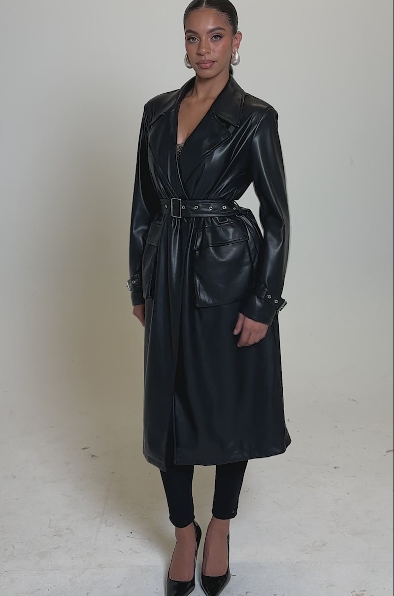 A video of a woman in the MONTANNA BUTTER LUXE EVERYDAY TRENCH, a long black vegan leather coat belted over black leggings and pointed heels, her hair sleekly pulled back and finished with hoop earrings.