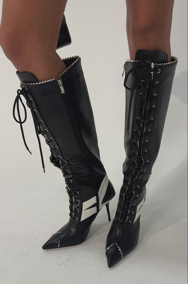 Video of woman modeling the AZALEA WANG JETER BLACK SPORTY STILETTO BOOTS with white stripes, holding a small black buckled handbag. The look is edgy and fashion-forward.