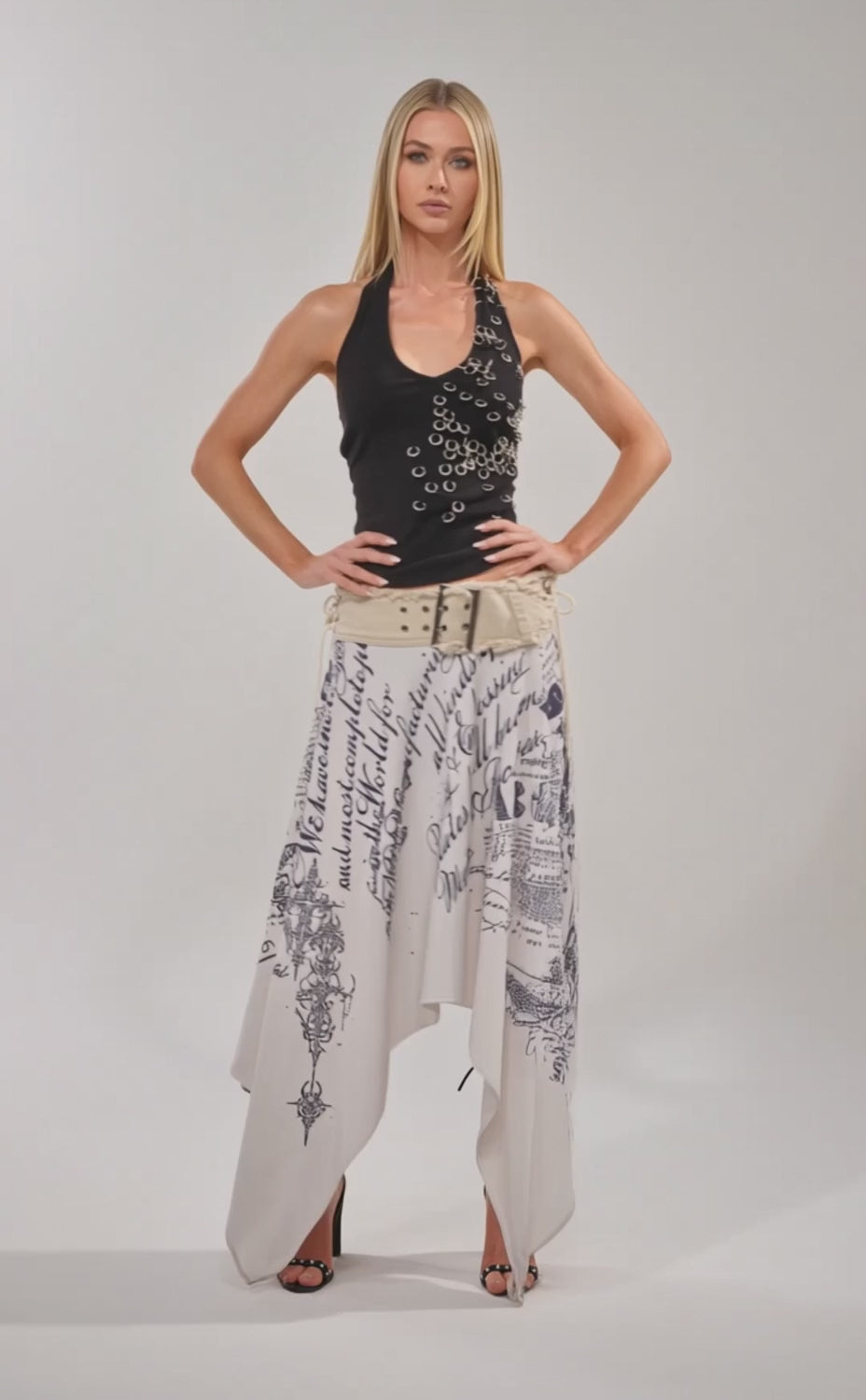 Video of woman modeling the RING RING RING EMBELLISHED TANK IN BLACK, paired with a studded shoulder bag, beige belt, and white script-print skirt. She poses confidently against a plain backdrop.