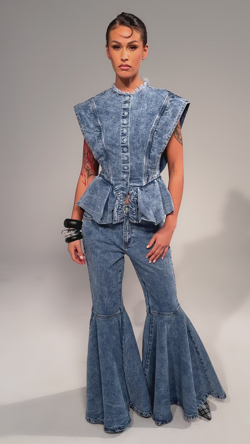 Video of woman modeling the DONT PLAY WITH MY HEART BELL BOTTOM DENIM, featuring flared jeans and a sleeveless, structured top. Tattoos cover one arm and chunky bracelets complete the look against a plain light gray background.