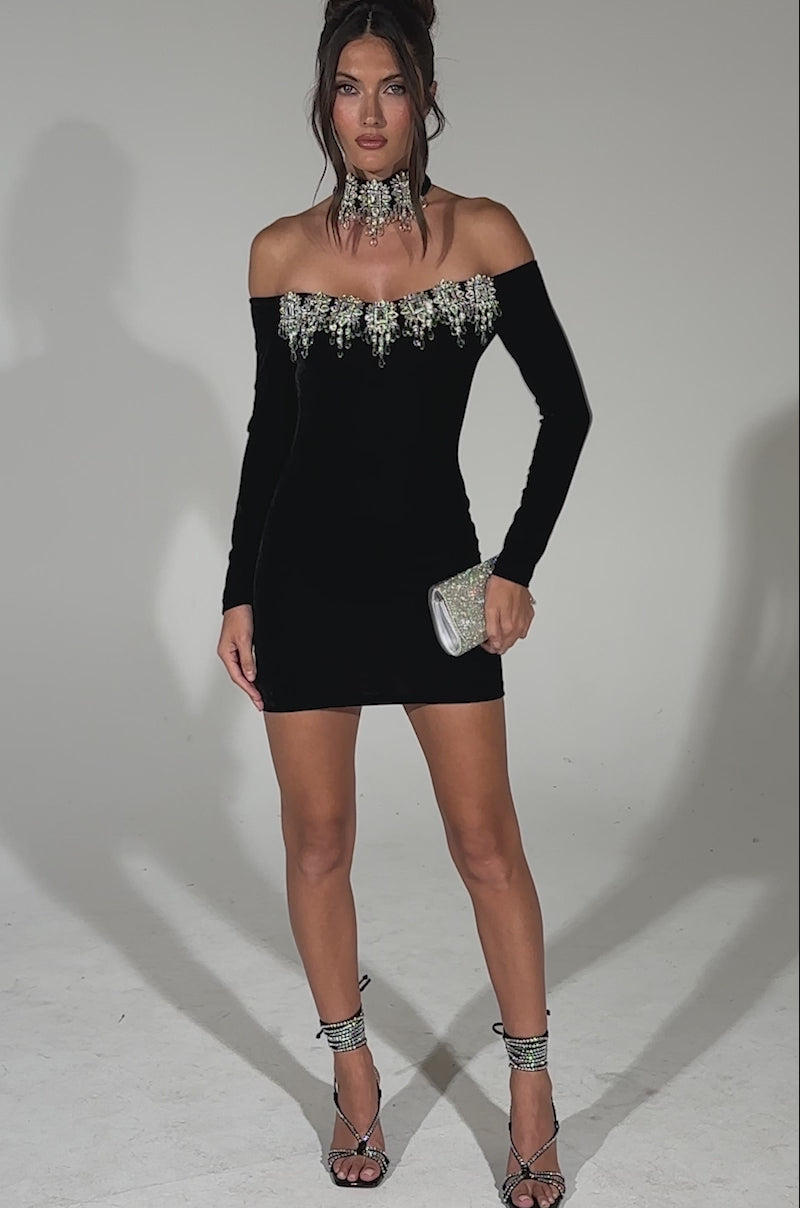 A video of a woman modeling the STAR OF THE NIGHT RHINESTONE MINI DRESS, a black velvet off-the-shoulder piece with silver embellishments, paired with statement jewelry, strappy heels, and a silver clutch against a plain light background.