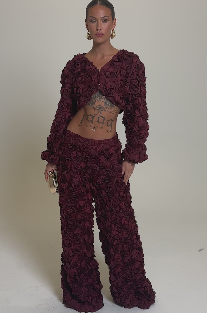 Video of woman modeling the ROSE PETALS FLORAL SHRUG—a burgundy cropped outfit with 3D roses—paired with gold hoop earrings, a gold textured clutch, slicked-back hair, and a striking “1999” tattoo across her chest and stomach.