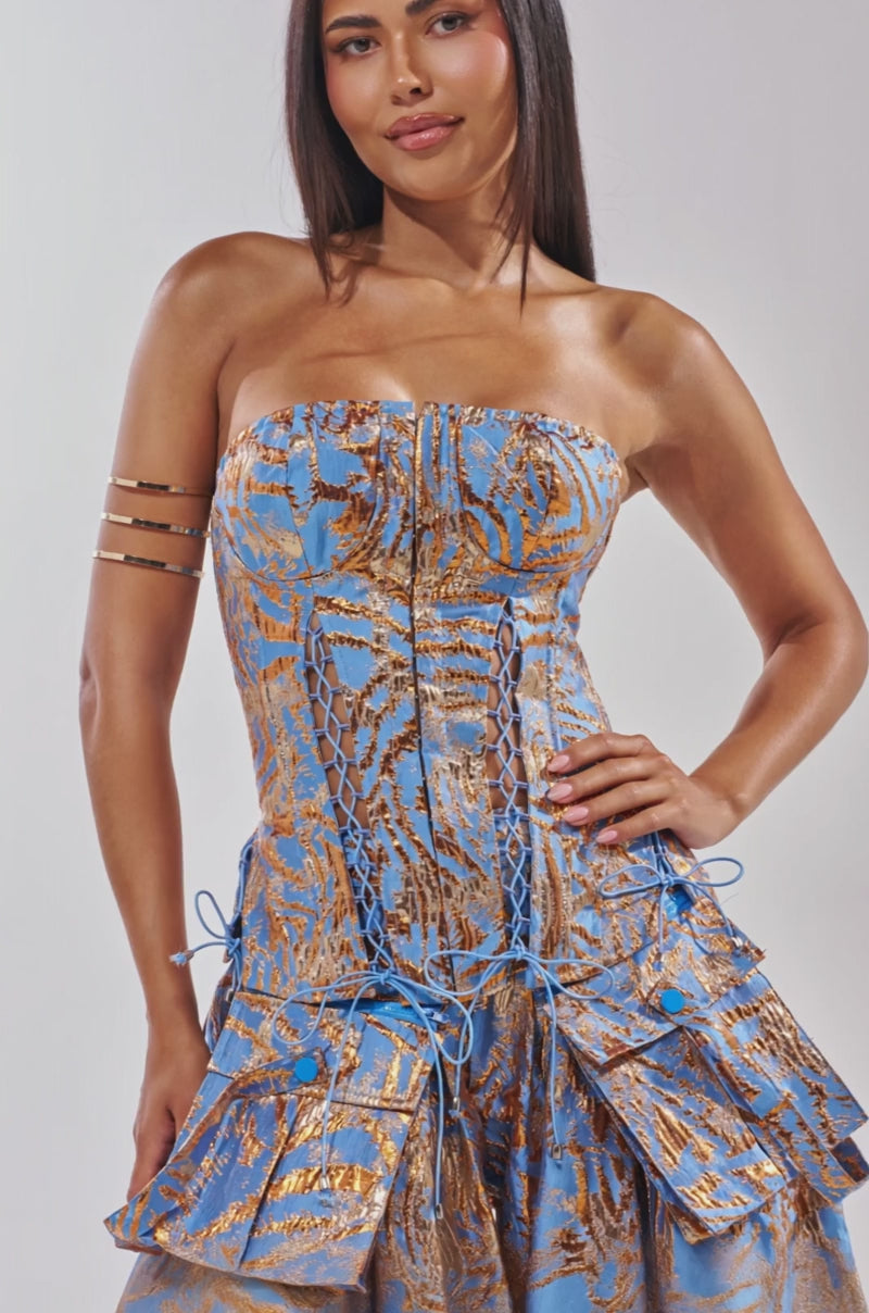 Video of woman modeling the WAYS TO GO BROCADE CORSET, a strapless blue and gold piece with metallic detailing. She stands smiling softly, one hand on her hip and gold arm cuffs adorning her upper arm.