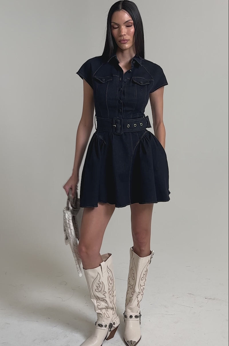 A video of a woman wearing the SADIE BUTTON UP DENIM MINI DRESS with a belted waist and white cowboy boots holds a small handbag adorned with a scarf. 