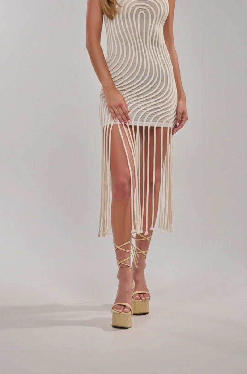 Video of woman modeling the ILLUSIONS SLEEVELESS MINI DRESS IN BONE, featuring a fitted, sleeveless silhouette with swirling textures and a rope fringe hem, styled with lace-up shoes for an effortlessly chic vacation look against a neutral backdrop.
