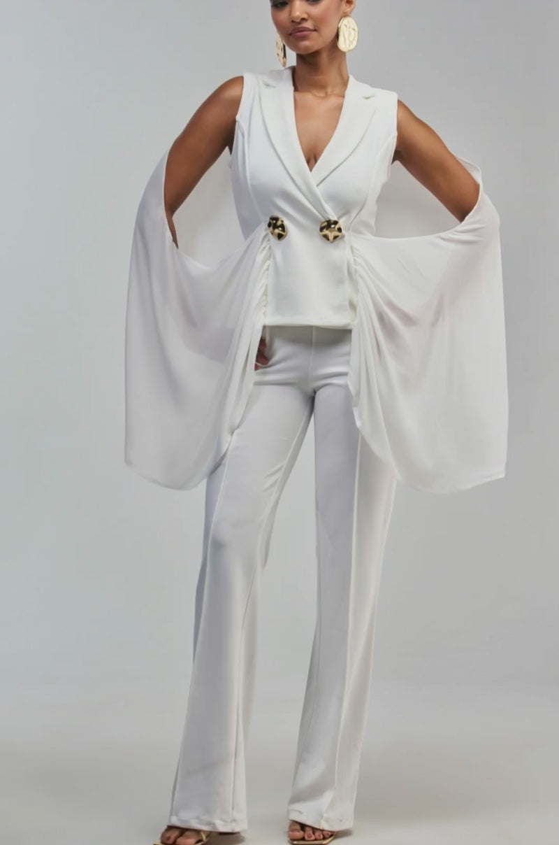 Video of a woman modeling the SHE IS THE MOMENT COLLARED JUMPSUIT, featuring a crisp white color, unique draped cape-like sleeves, and sophisticated gold-button accents.