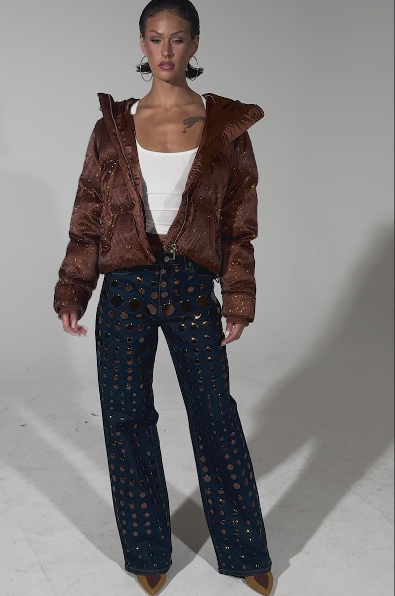 A video of a person wearing the FAYE RHINESTONE PUFFER IN COPPER over a white tank top and navy blue pants with large circular cutouts, standing against a plain background and looking confidently to the side.