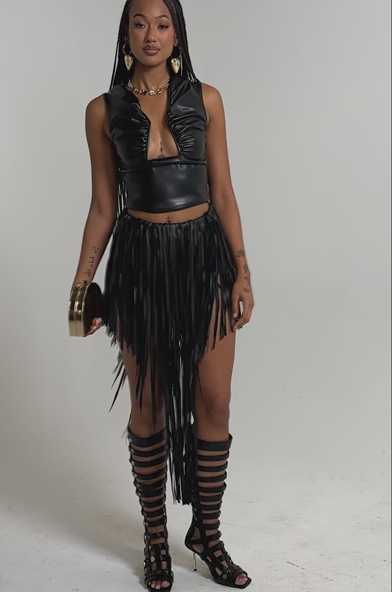 A video of a woman in a black leather sleeveless crop top with a front cutout, the COACHELLA FRINGE SKIRT, gladiator-style knee-high sandals, gold earrings, and holding a small round clutch.