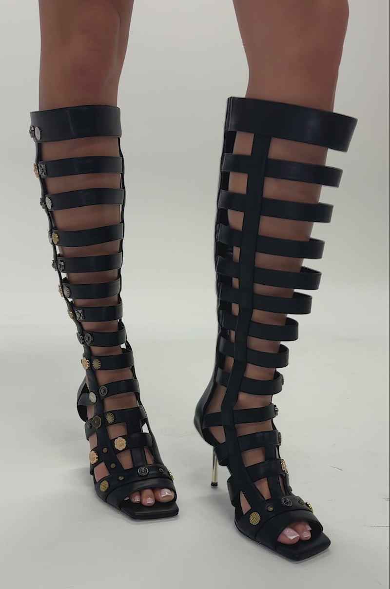 Video of woman modeling the AZALEA WANG GOLD WENTWORTH Gladiator Open Toe Sandals featuring black knee-high straps and two-toned hardware embellishments, including gold coins, standing on a light gray surface. The stiletto heel adds extra elegance.