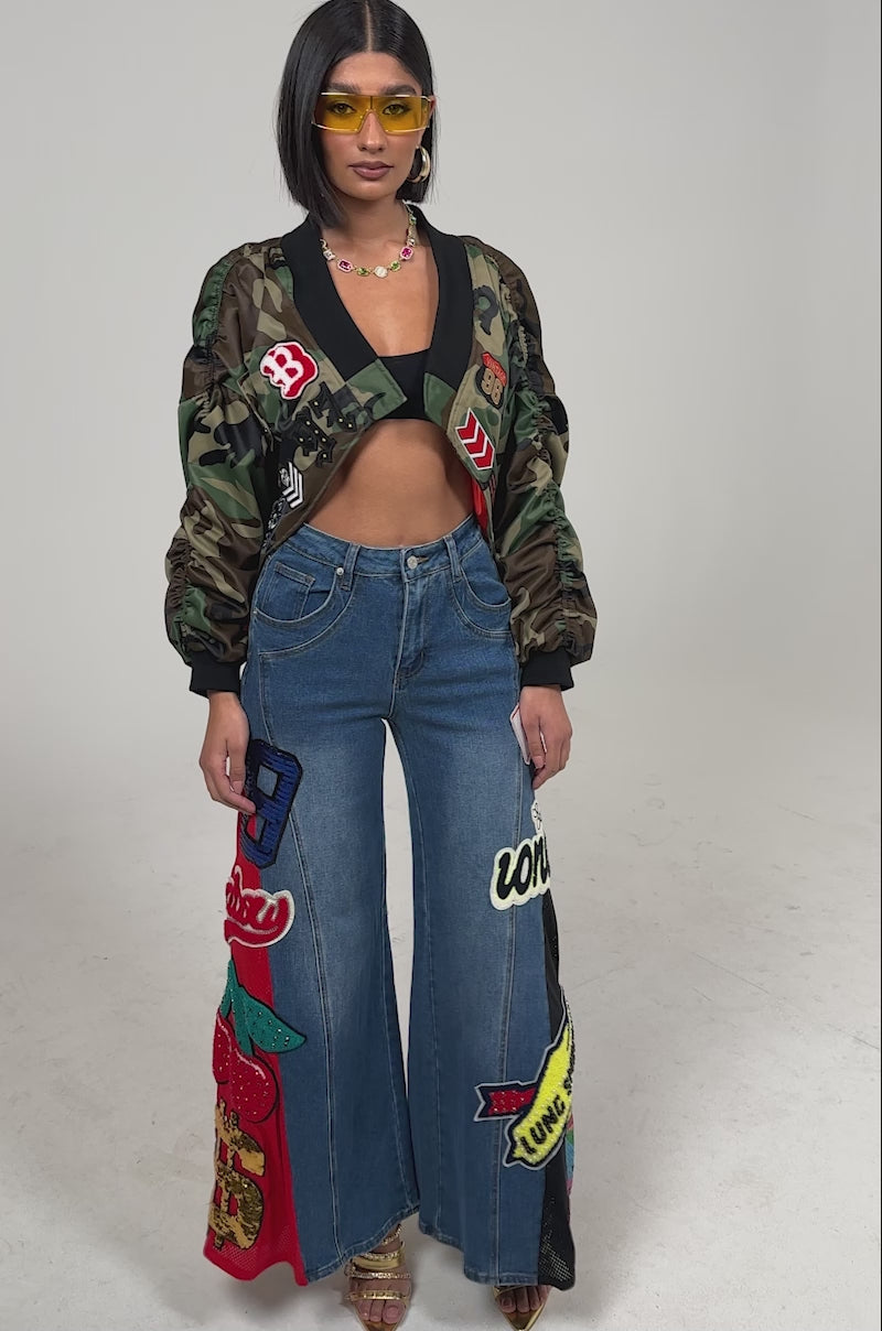 Video of woman modeling the NOT SORRY PATCHWORK DENIM JEANS with colorful designs, gold high-heeled sandals, and a cropped multi-patterned jacket.