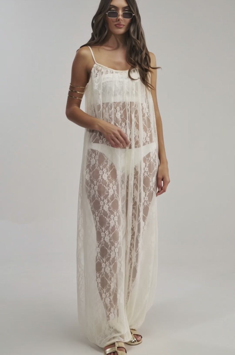 A video of a woman in the MYSTICAL BLOOM LACE JUMPSUIT over white underwear, paired with gold bracelets, sandals, and sunglasses against a plain light backdrop.