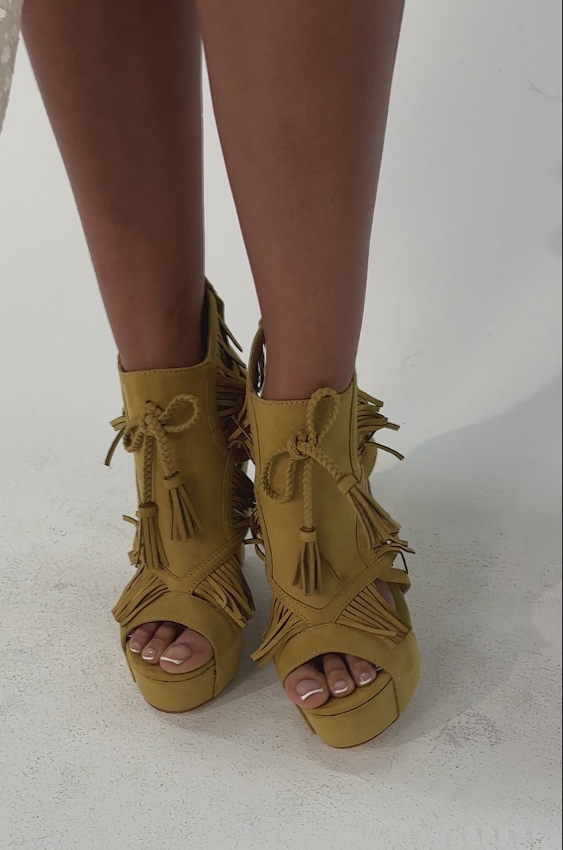 Video of woman modeling the AZALEA WANG LEONTINE WARM OLIVE WEDGE FAUX SUEDE SANDAL, featuring fringe details, cutouts, open toes. The stylish platform wedge heels stand out against a plain light background.