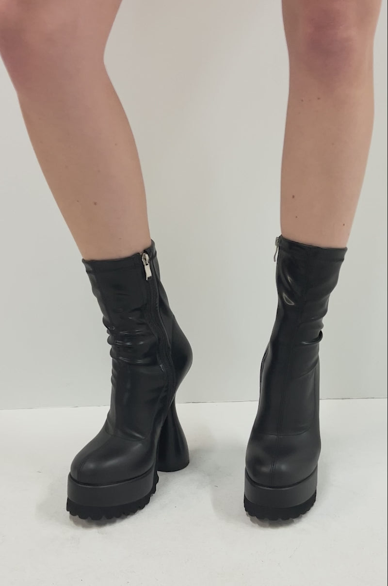 Video of person wearing the AZALEA WANG WARIO BLACK CHUNKY BOOTIE—black, high-heeled faux leather booties with a sculpted heel and shiny, fitted design