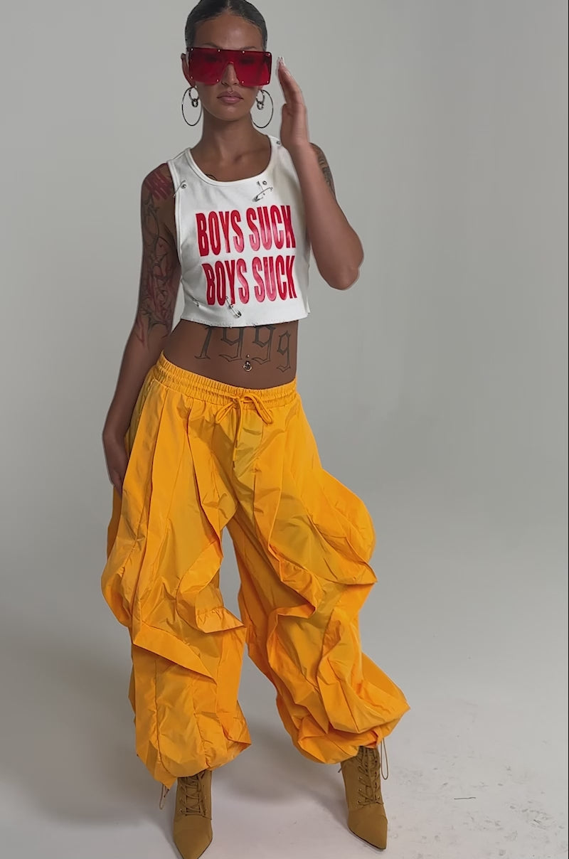 A video of a person wearing a white "BOYS SUCK" crop top, HOTTEST ON THE BLOCK JOGGER bright yellow pants, tan boots, and large hoop earrings.
