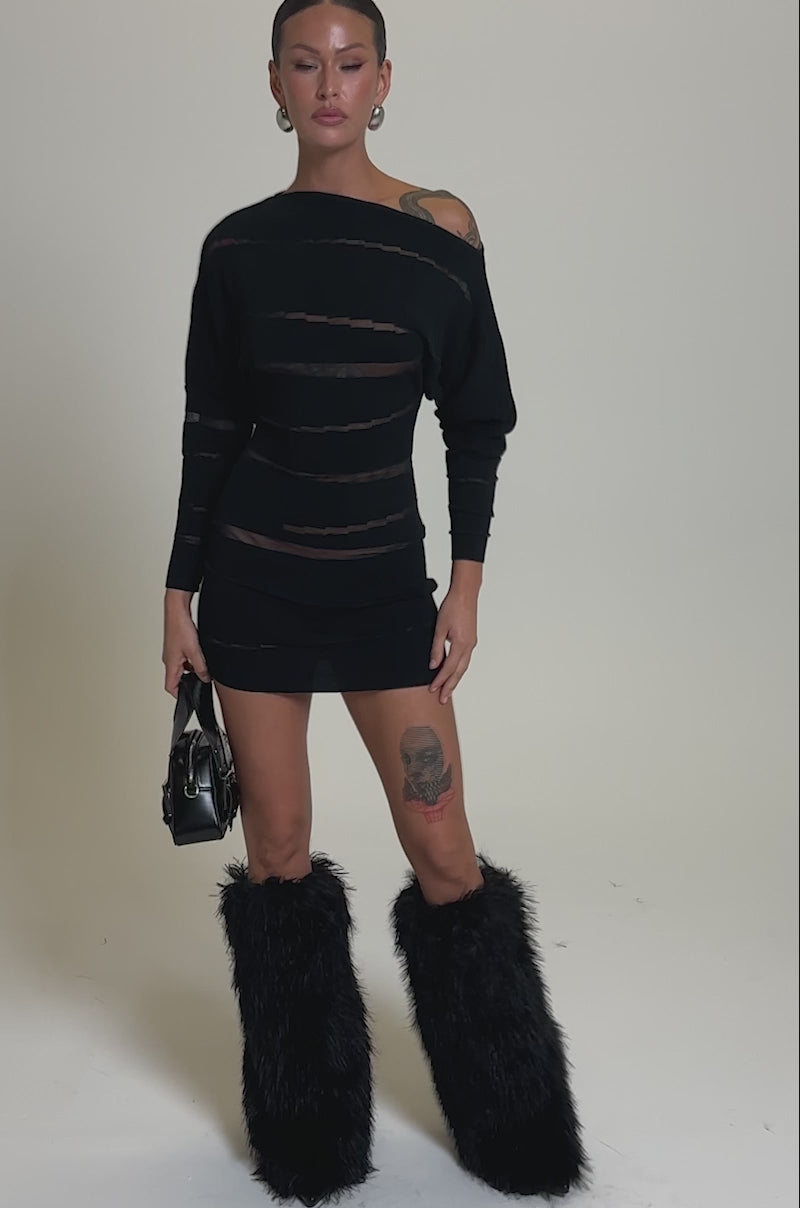A video of a woman against a plain background in the AFTER DARK STRIPED MINI DRESS featuring curve-hugging black mesh stripes, paired with black furry knee-high boots, a small black handbag, hoop earrings, slicked-back hair, and visible tattoos.