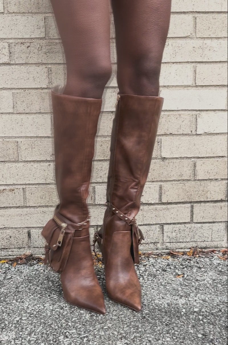 Video of woman modeling the AZALEA WANG RONDEL BROWN DISTRESSED STILETTO BOOT with gold details and a small pouch stands against a light brick wall.