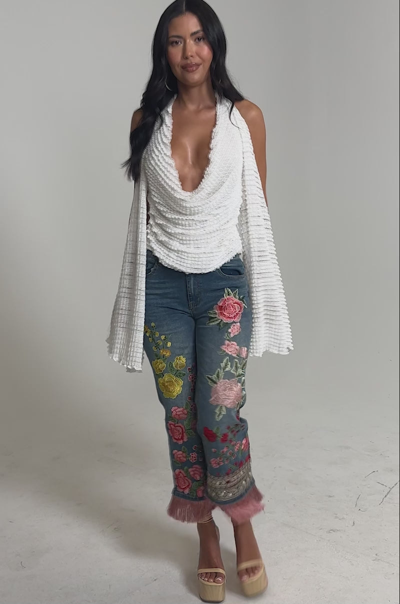 Video of woman modeling the TAKE ME TO THE TROPICS COWL NECK BLOUSE, a white crochet halter top with a deep cowl neckline, paired with blue jeans featuring colorful floral embroidery against a plain light background.