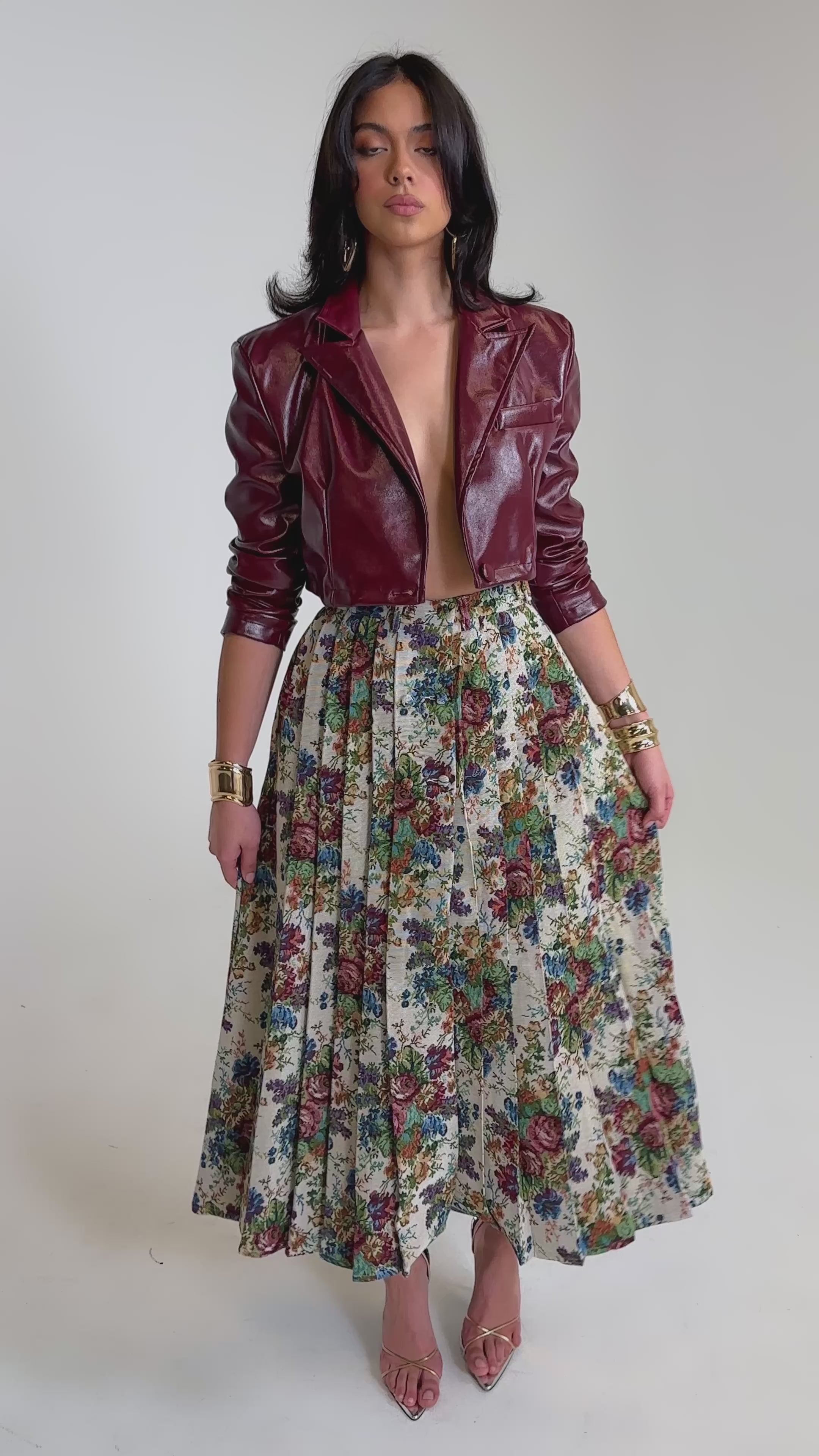 A video of a woman in the TIFFANY BROCADE MAXI SKIRT paired with gold jewelry, strappy high heels, and a burgundy faux leather jacket against a plain light background.