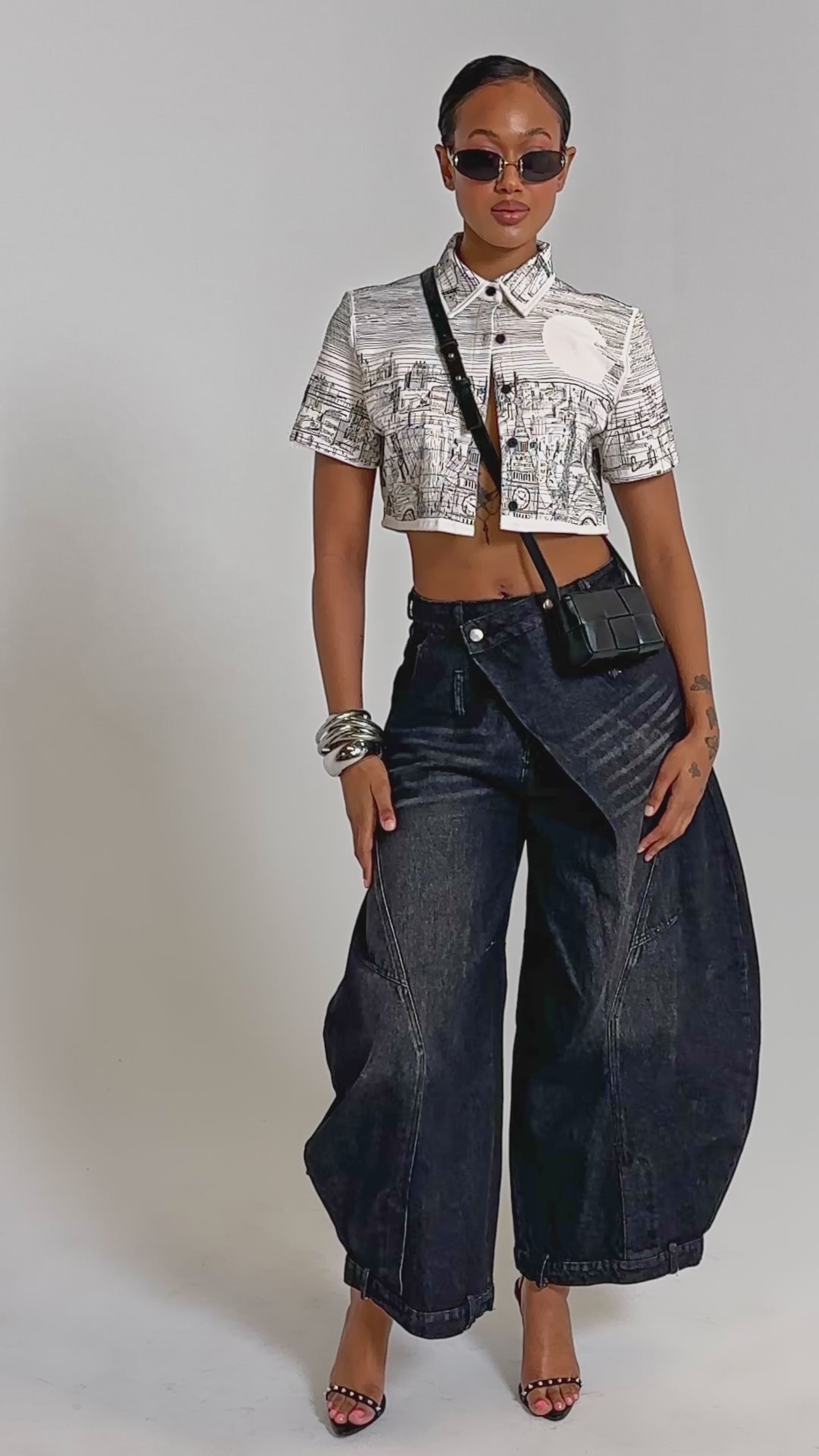 A video of a woman wearing the INK HORIZON EMBROIDERED TOP, wide-leg black jeans, high heels, and a black crossbody bag, accessorized with bracelets and hoop earrings.